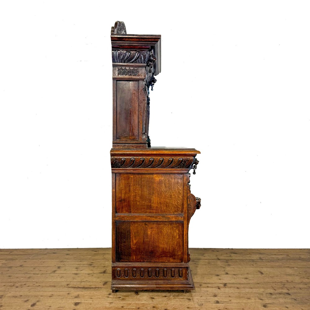 A two-tier dark oak cupboard featuring relief carvings, lion masks, geometric panelled doors, and a decorative carved pediment. View XI