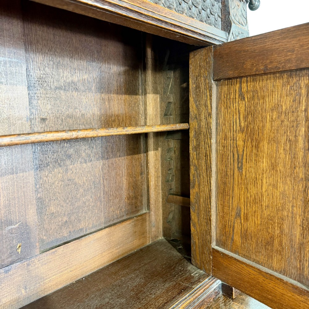 A two-tier dark oak cupboard featuring relief carvings, lion masks, geometric panelled doors, and a decorative carved pediment. View XII