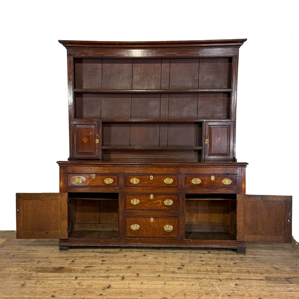 An antique Georgian dark oak dresser featuring an upper shelving rack, small cupboard doors with diamond inlay, and a base containing multiple drawers and panelled cupboards with brass handles. View II