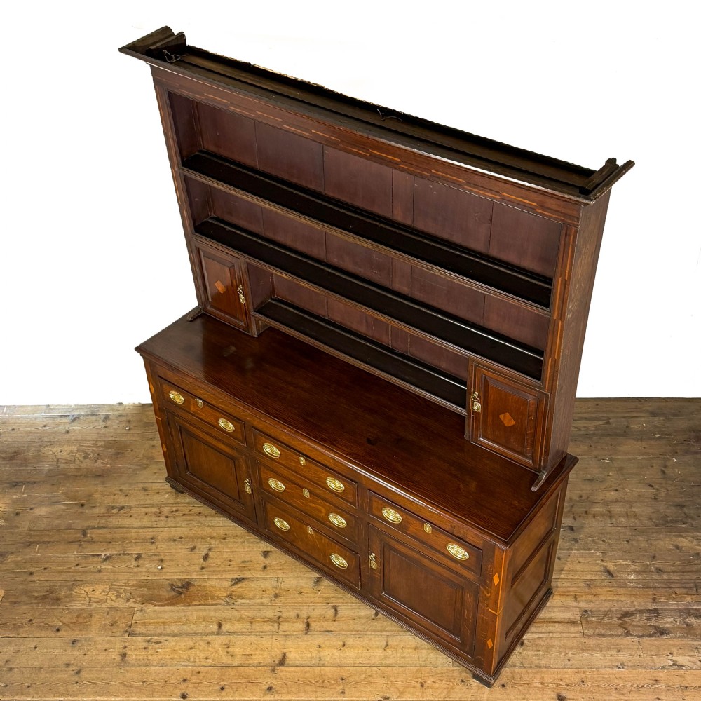 An antique Georgian dark oak dresser featuring an upper shelving rack, small cupboard doors with diamond inlay, and a base containing multiple drawers and panelled cupboards with brass handles. View III