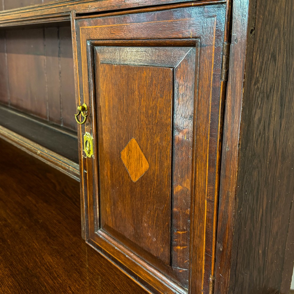 An antique Georgian dark oak dresser featuring an upper shelving rack, small cupboard doors with diamond inlay, and a base containing multiple drawers and panelled cupboards with brass handles. View V