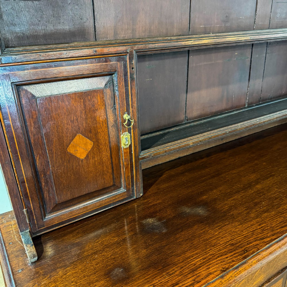 An antique Georgian dark oak dresser featuring an upper shelving rack, small cupboard doors with diamond inlay, and a base containing multiple drawers and panelled cupboards with brass handles. View IX