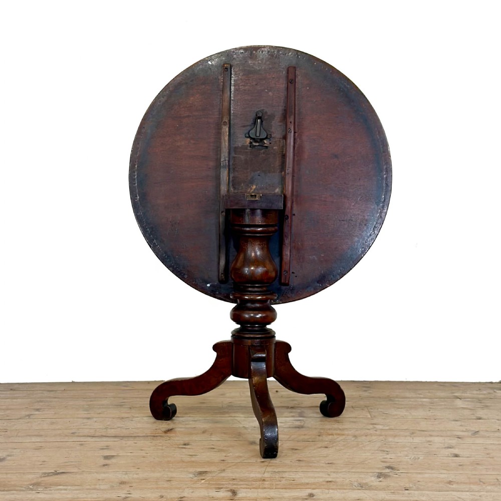 An antique Georgian mahogany tilt top table shown upright with a circular top, turned central pedestal, and tripod legs. View IV