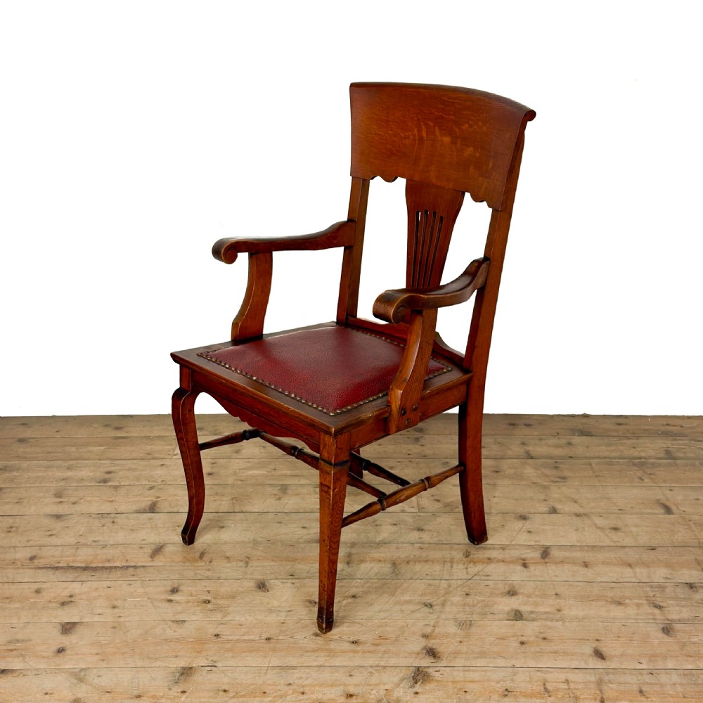 An antique dark oak carver chair featuring a carved splat backrest, curved armrests, turned stretchers, and a padded red leather seat with brass studs.
