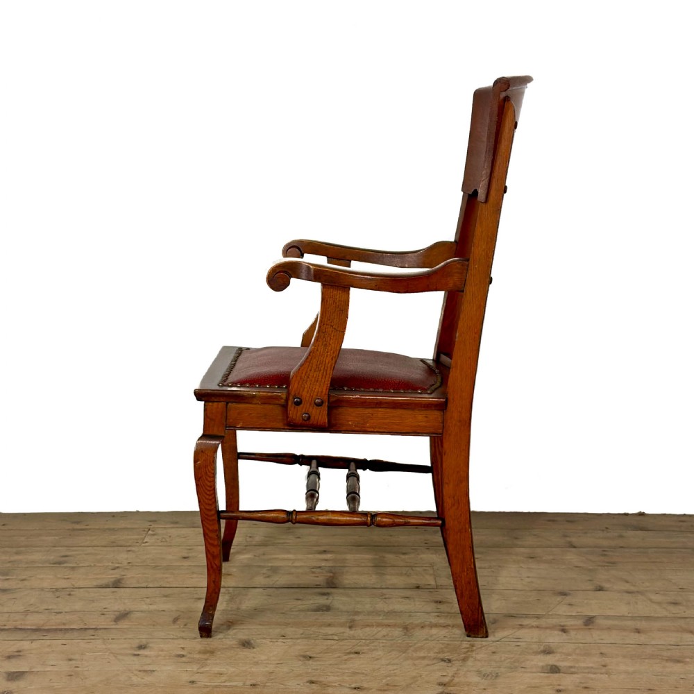 An antique dark oak carver chair featuring a carved splat backrest, curved armrests, turned stretchers, and a padded red leather seat with brass studs. View II