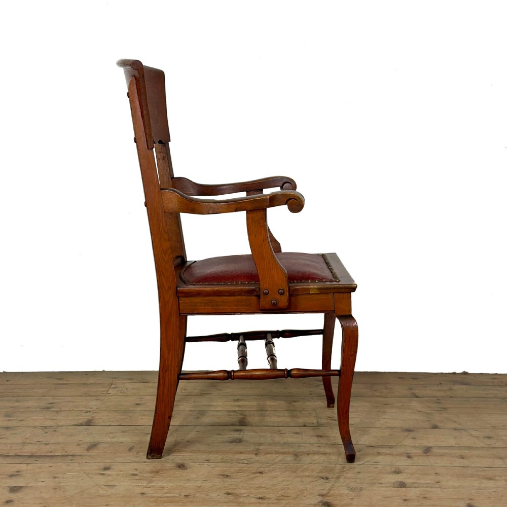An antique dark oak carver chair featuring a carved splat backrest, curved armrests, turned stretchers, and a padded red leather seat with brass studs. View V