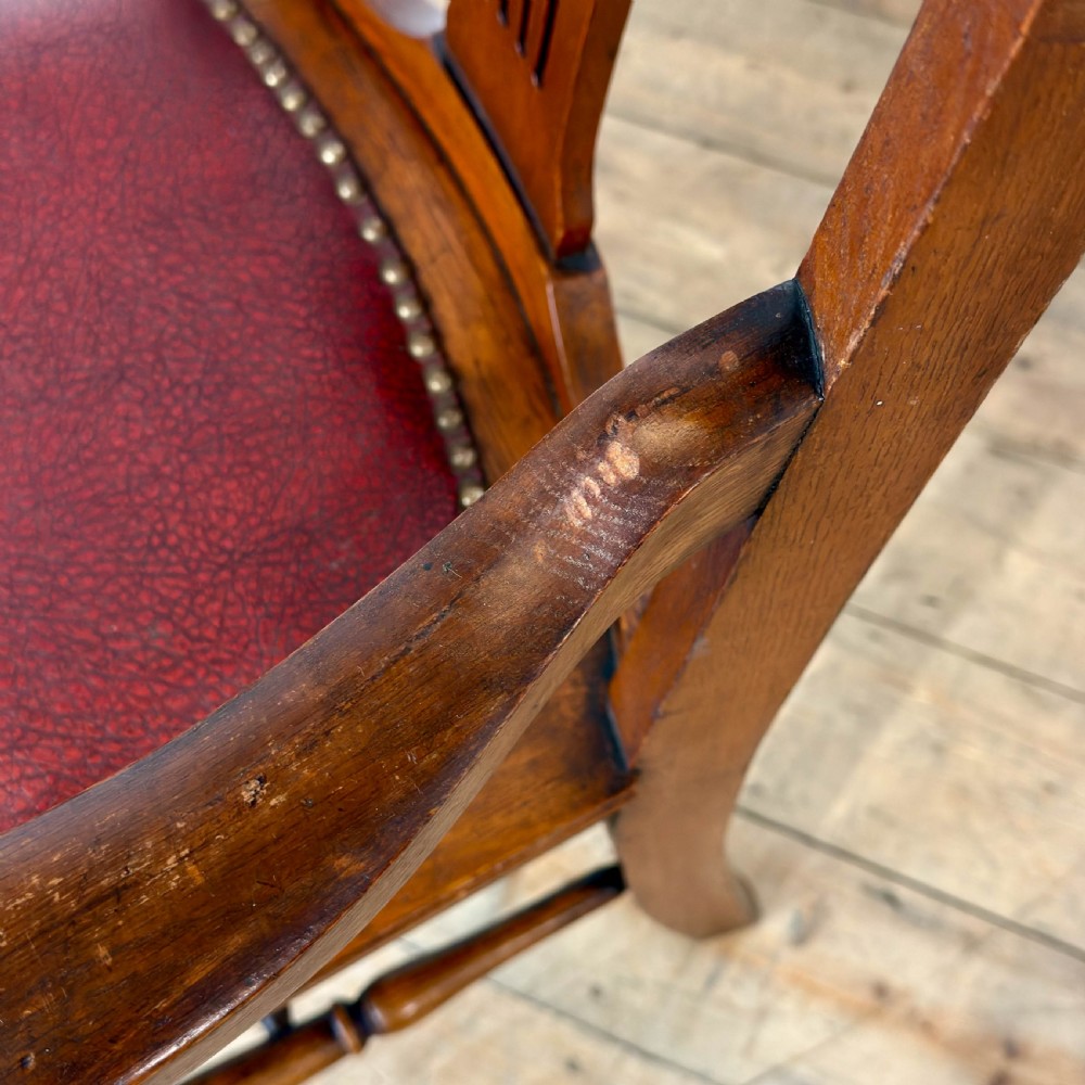 An antique dark oak carver chair featuring a carved splat backrest, curved armrests, turned stretchers, and a padded red leather seat with brass studs. View VI