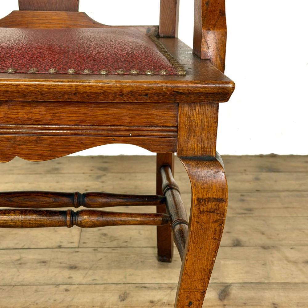 An antique dark oak carver chair featuring a carved splat backrest, curved armrests, turned stretchers, and a padded red leather seat with brass studs. View VII