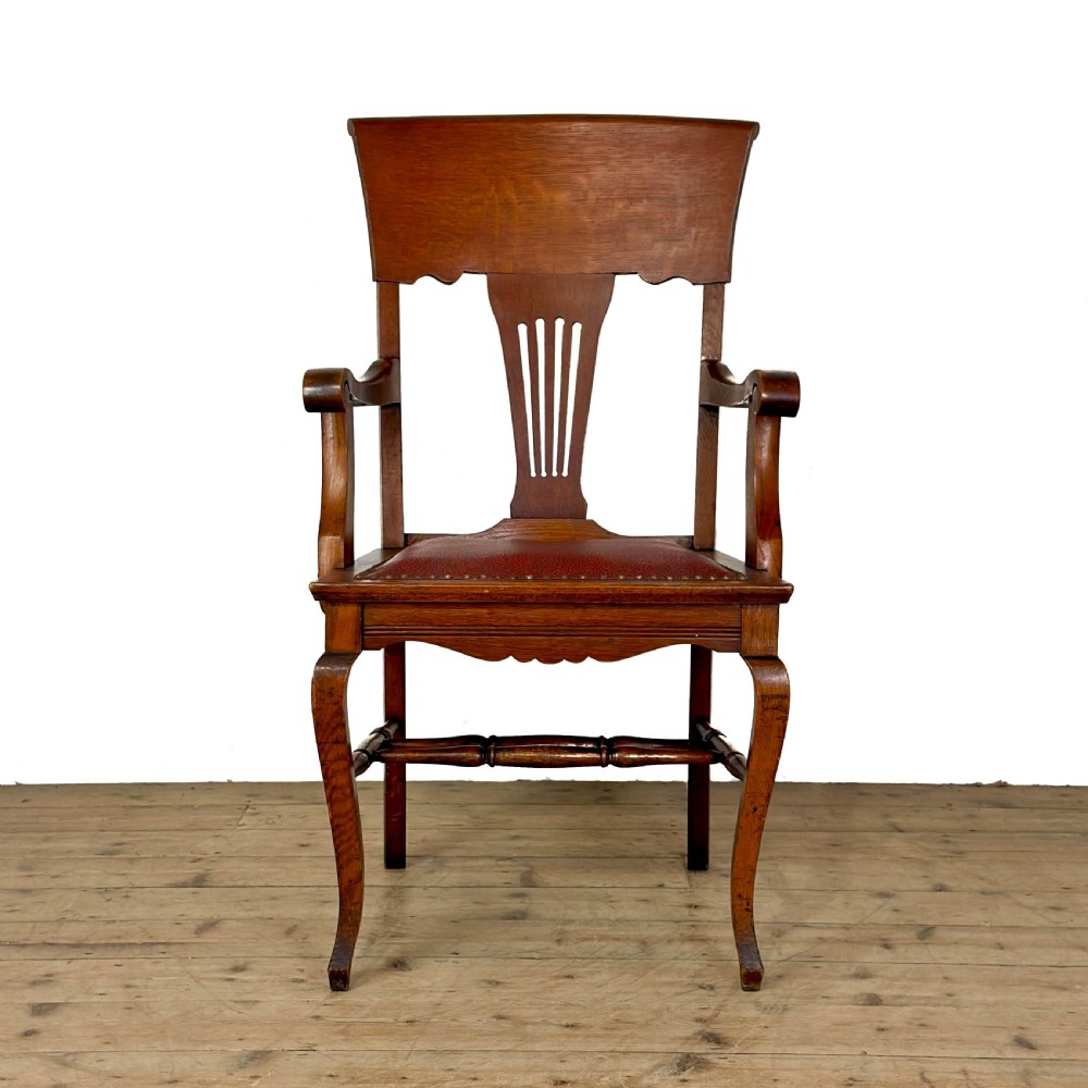 An antique dark oak carver chair featuring a carved splat backrest, curved armrests, turned stretchers, and a padded red leather seat with brass studs. View IX