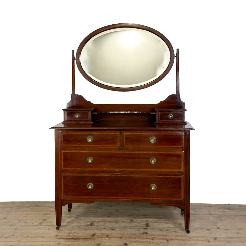 An Edwardian mahogany dressing table featuring an oval swivel mirror, satinwood stringing inlay, multiple drawers with brass handles, and square tapered legs on castors.