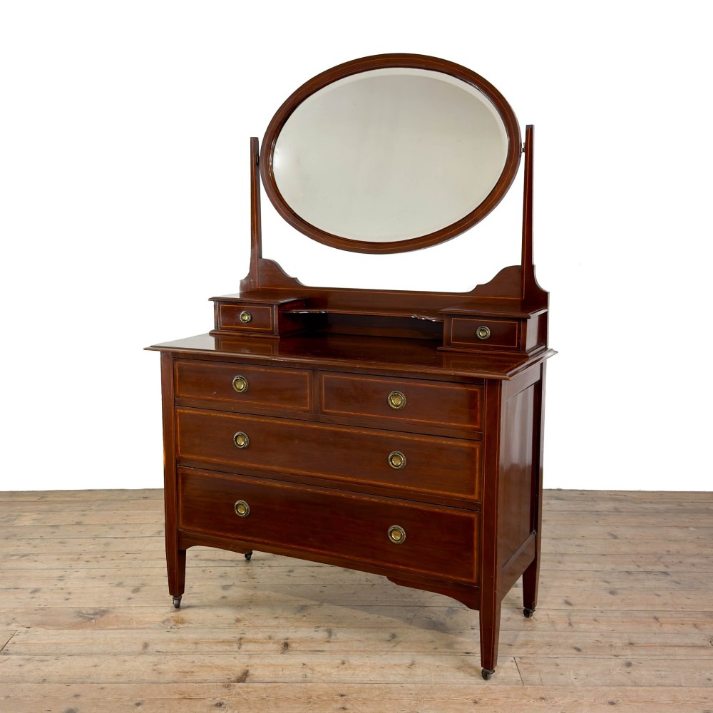 An Edwardian mahogany dressing table featuring an oval swivel mirror, satinwood stringing inlay, multiple drawers with brass handles, and square tapered legs on castors. View VI