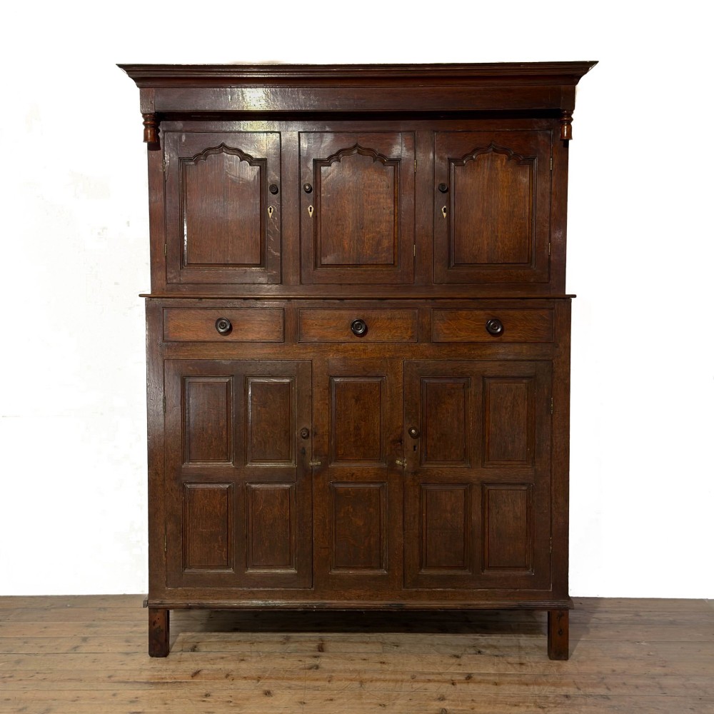 An 18th-century dark oak Welsh cwpwrdd deuddarn cupboard featuring two tiers with arched panelled doors and three lower drawers.