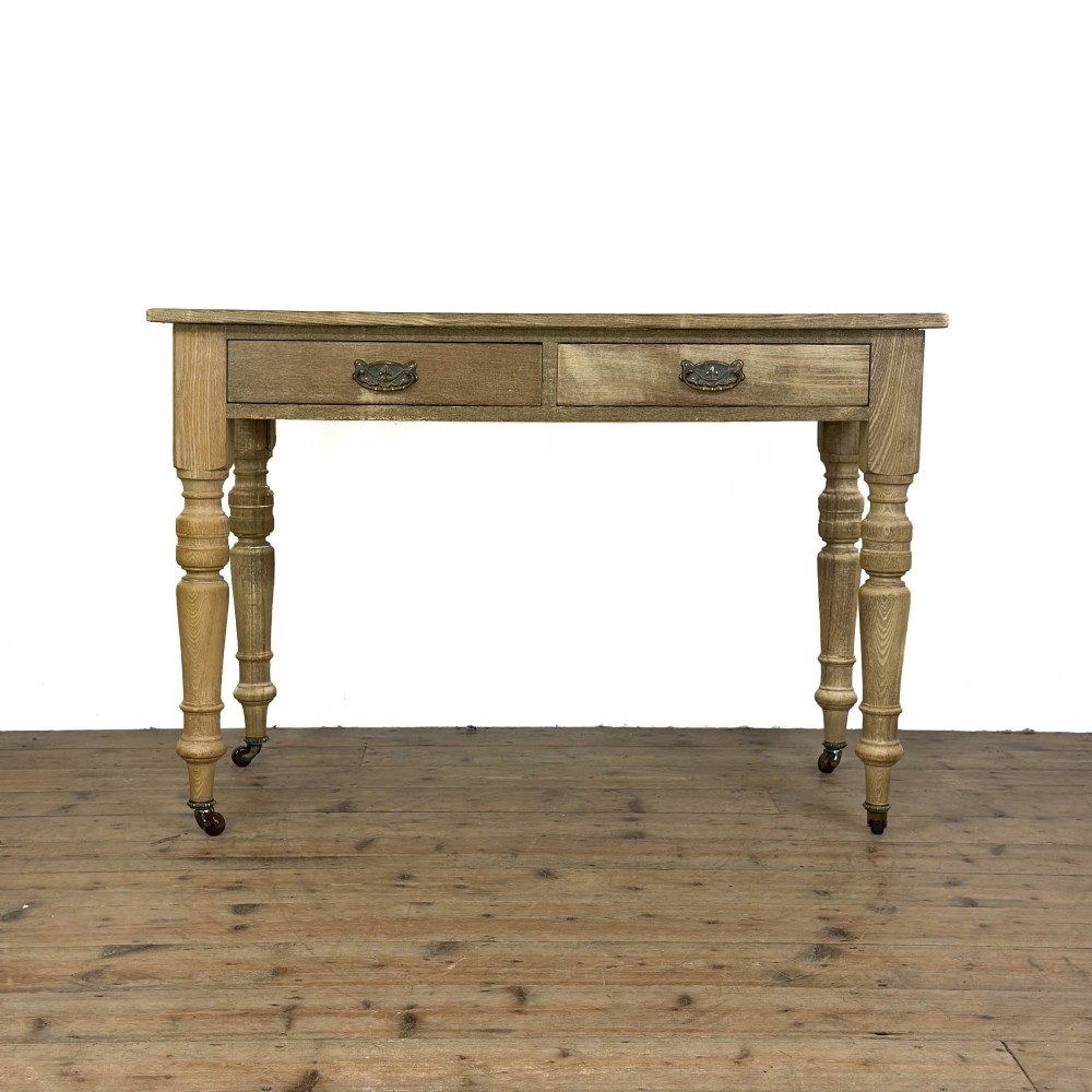 An antique stripped pine side table featuring two drawers with metal drop handles and four turned legs on castors.