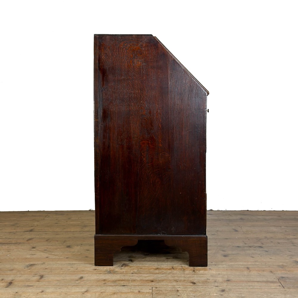 An antique Georgian oak writing bureau featuring a sloping fall-front lid above four drawers with brass swan-neck handles. View X