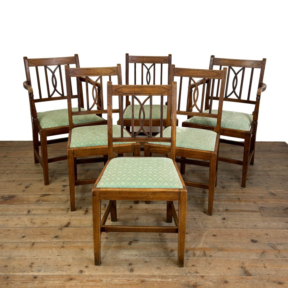 A set of six antique oak dining chairs, including two carvers, featuring geometric inlaid crest rails, decorative back splats, and light green patterned fabric seats.