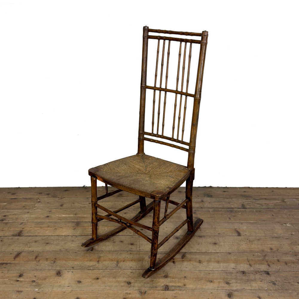 An antique dark brown wooden rocking chair with faux bamboo turnings, a high spindle back, and a woven rush seat.