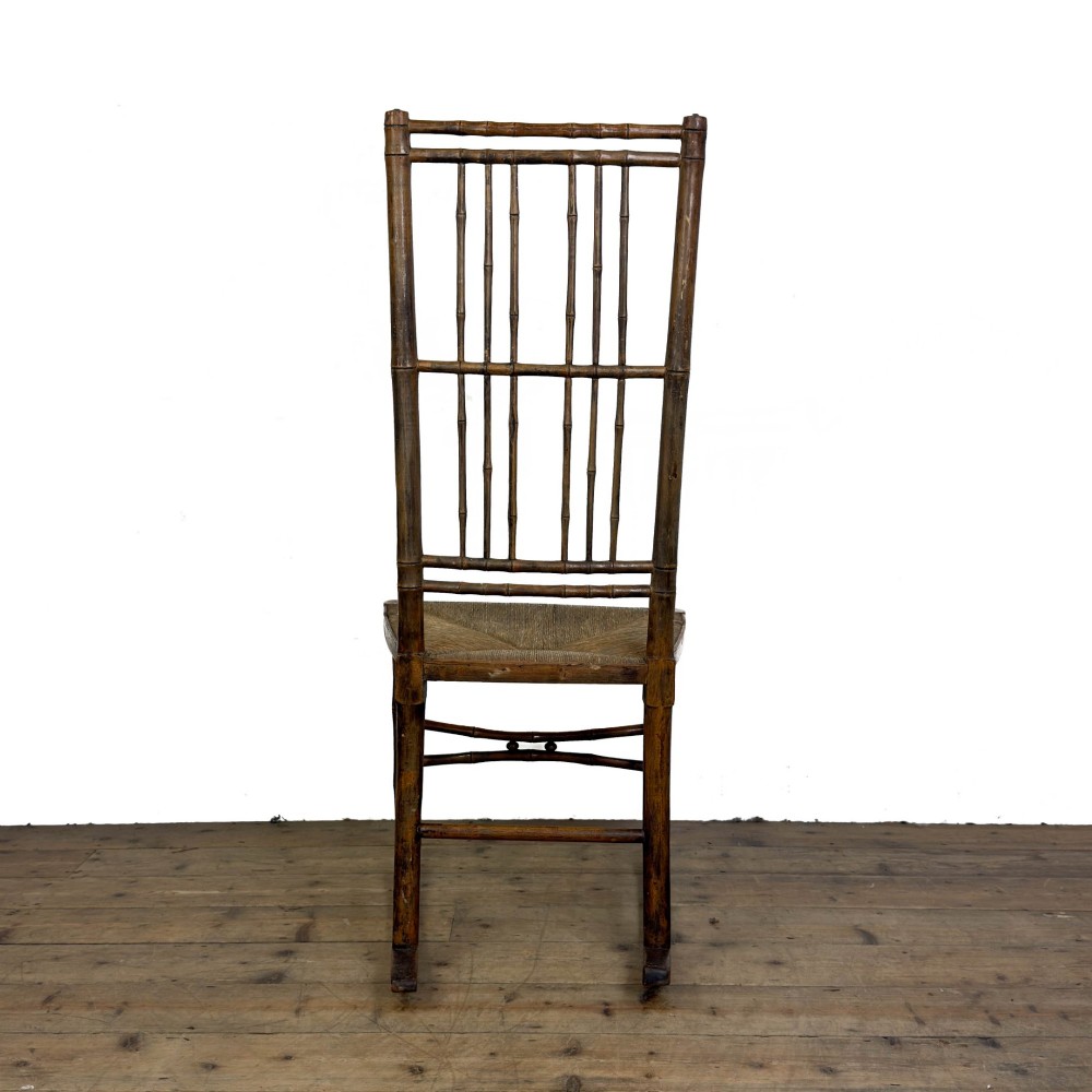 An antique dark brown wooden rocking chair with faux bamboo turnings, a high spindle back, and a woven rush seat. View V