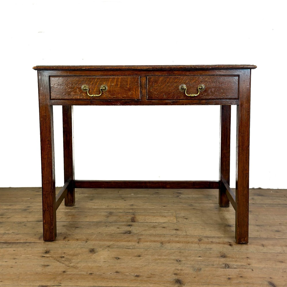 An antique dark oak writing table featuring two frieze drawers with brass drop handles, square legs, and low side stretchers.