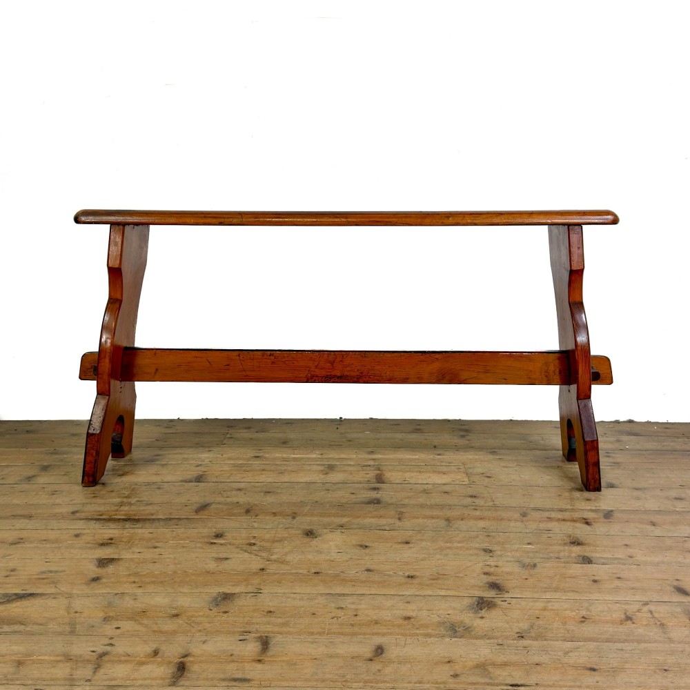 An antique pine bench featuring a long plank top, shaped trestle end supports, and a lower wooden stretcher rail.