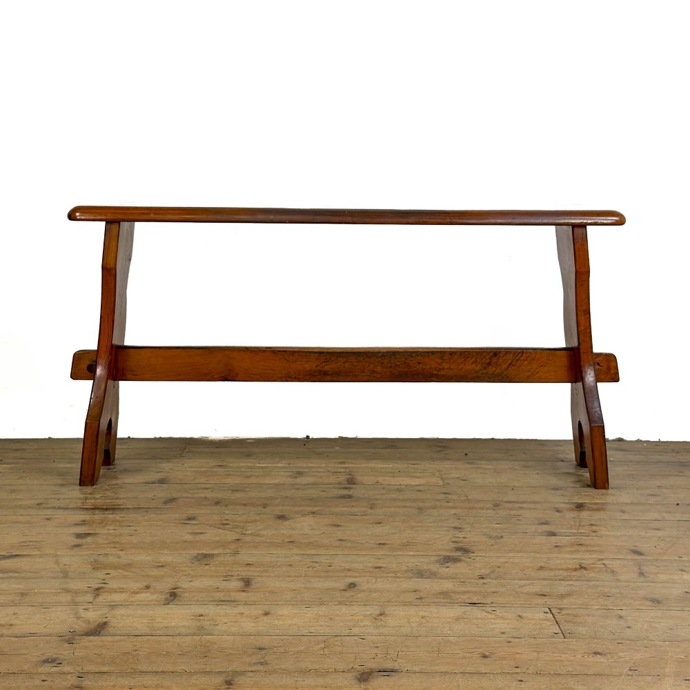 An antique pine bench featuring a long plank top, shaped trestle end supports, and a lower wooden stretcher rail. View VI