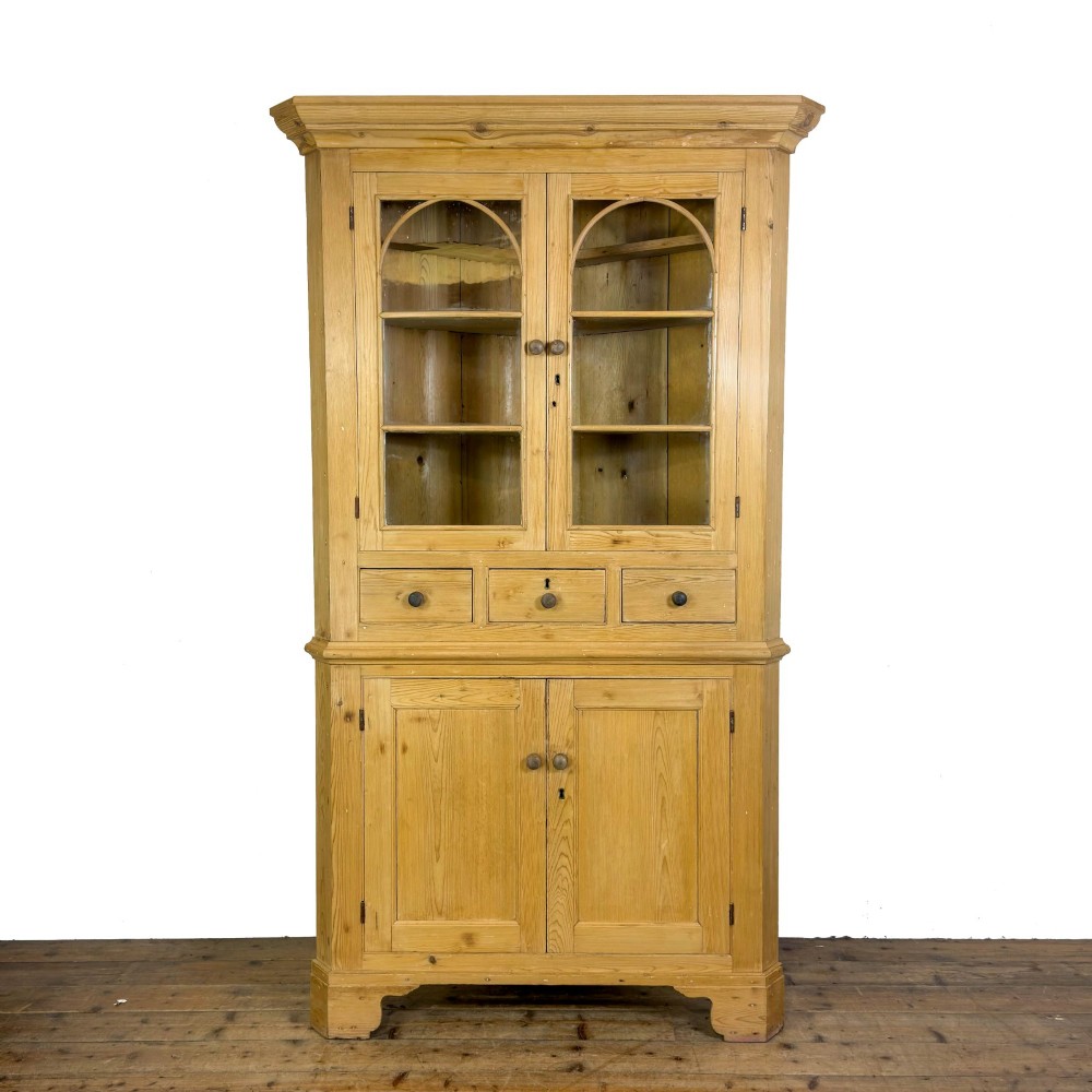 An antique stripped pine corner cabinet featuring arched glazed doors on the upper section, three drawers, and twin panelled cupboard doors below.