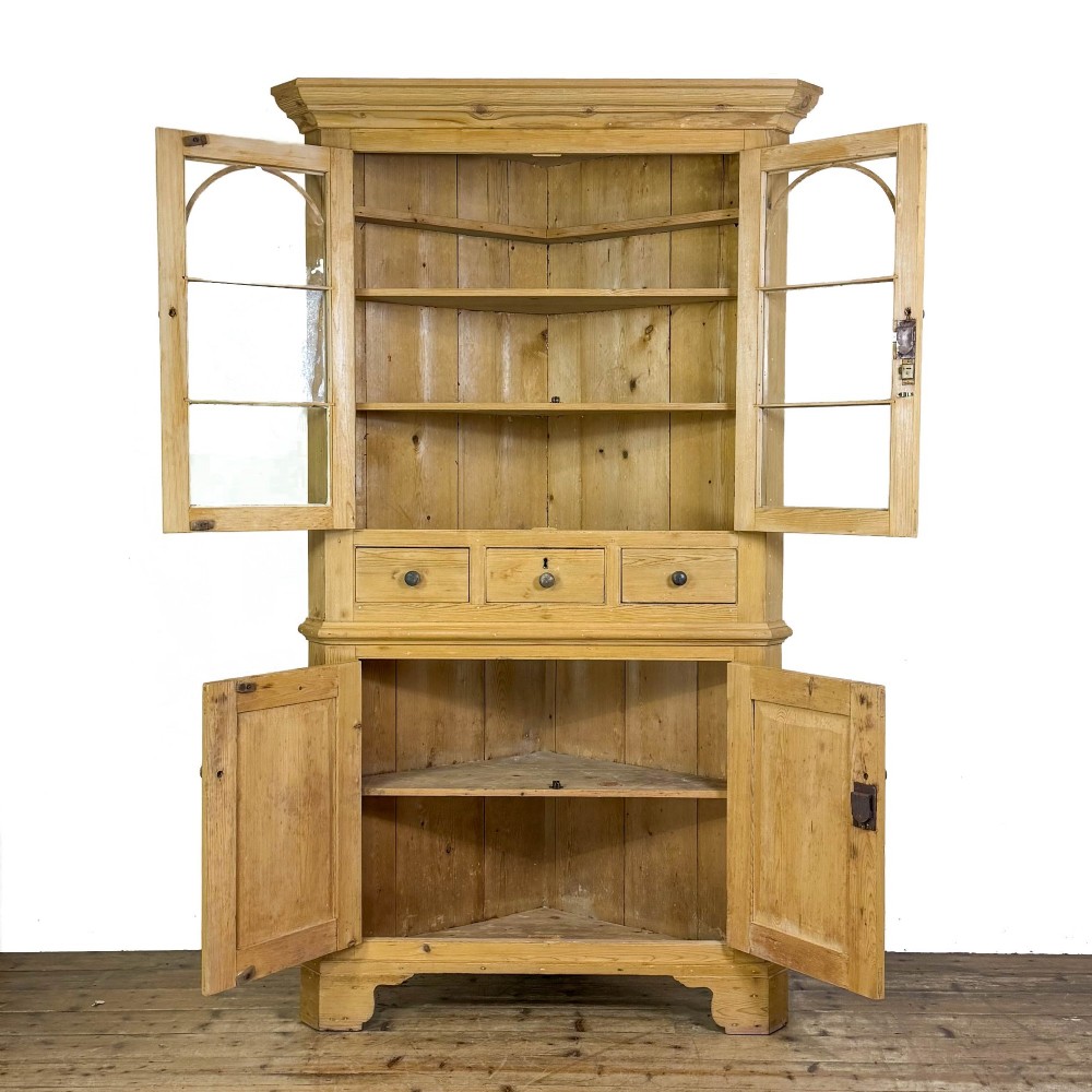 An antique stripped pine corner cabinet featuring arched glazed doors on the upper section, three drawers, and twin panelled cupboard doors below. View II