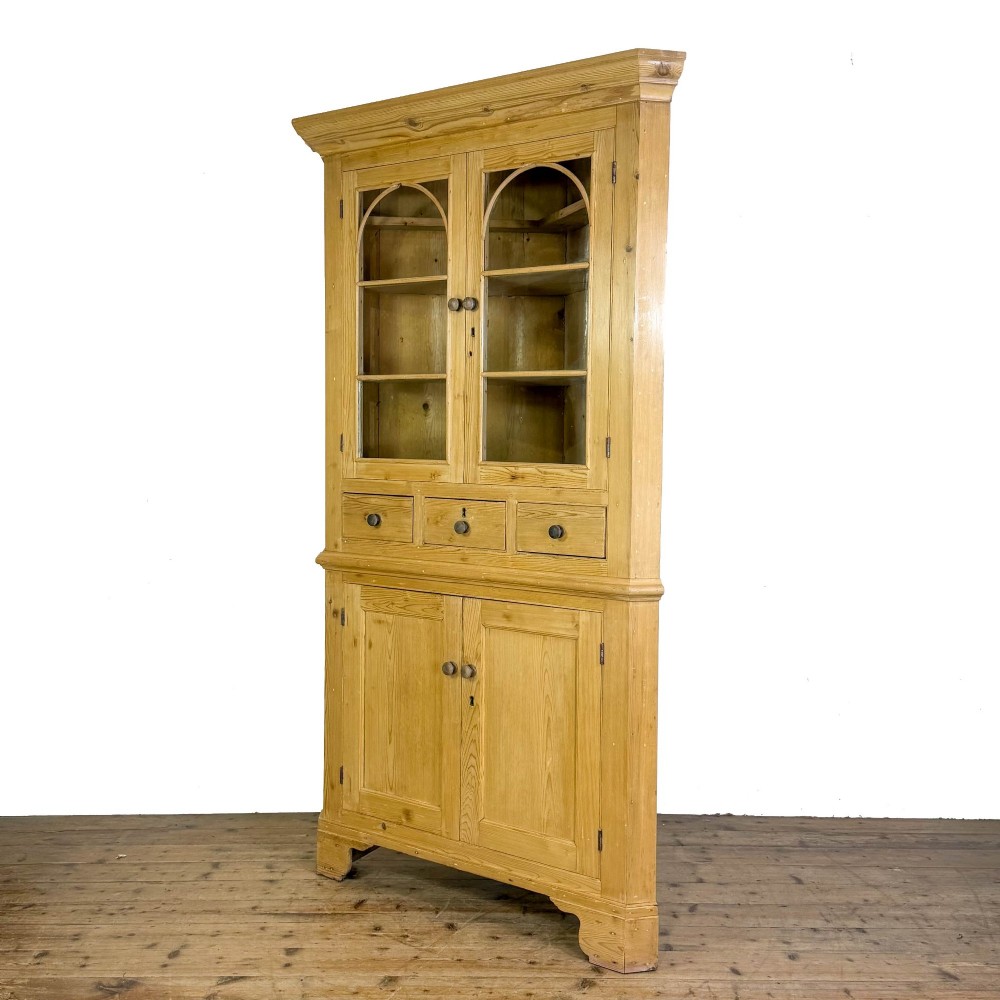 An antique stripped pine corner cabinet featuring arched glazed doors on the upper section, three drawers, and twin panelled cupboard doors below. View IV