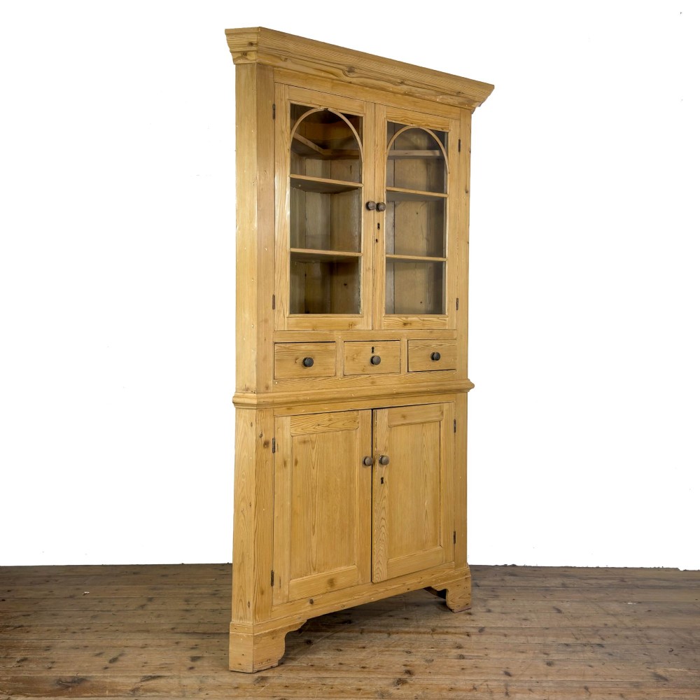 An antique stripped pine corner cabinet featuring arched glazed doors on the upper section, three drawers, and twin panelled cupboard doors below. View XI