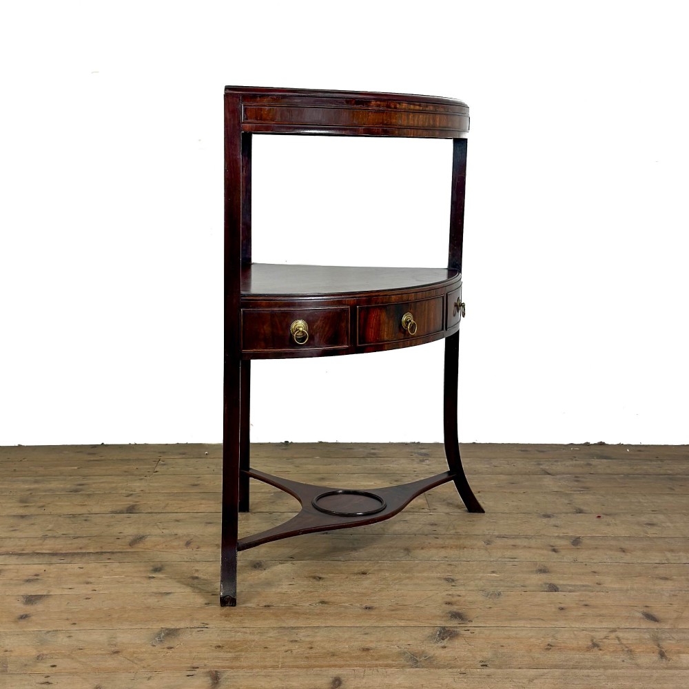 A Georgian dark mahogany triangular corner washstand featuring a smooth top shelf, a middle tier with three drawer fronts and brass ring handles, and slender legs connected by a lower stretcher. View VI