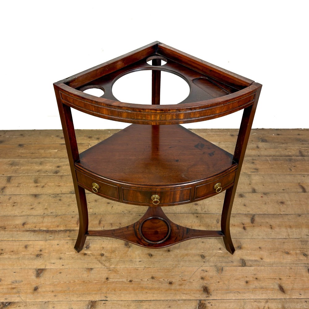 A Georgian dark mahogany triangular corner washstand featuring a smooth top shelf, a middle tier with three drawer fronts and brass ring handles, and slender legs connected by a lower stretcher. View X
