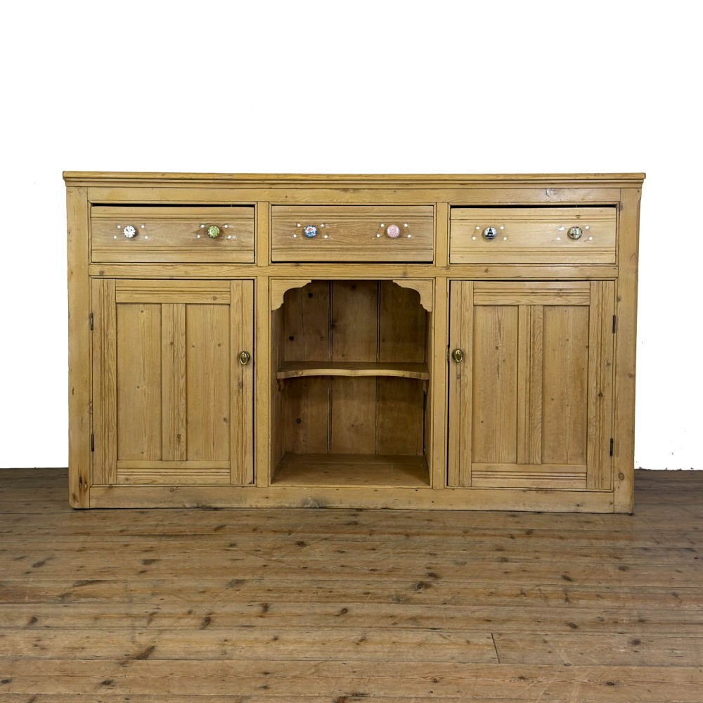 A Victorian pine dresser base with three frieze drawers, ceramic drawer knobs, two panelled cupboard doors, and a central open shelving recess.