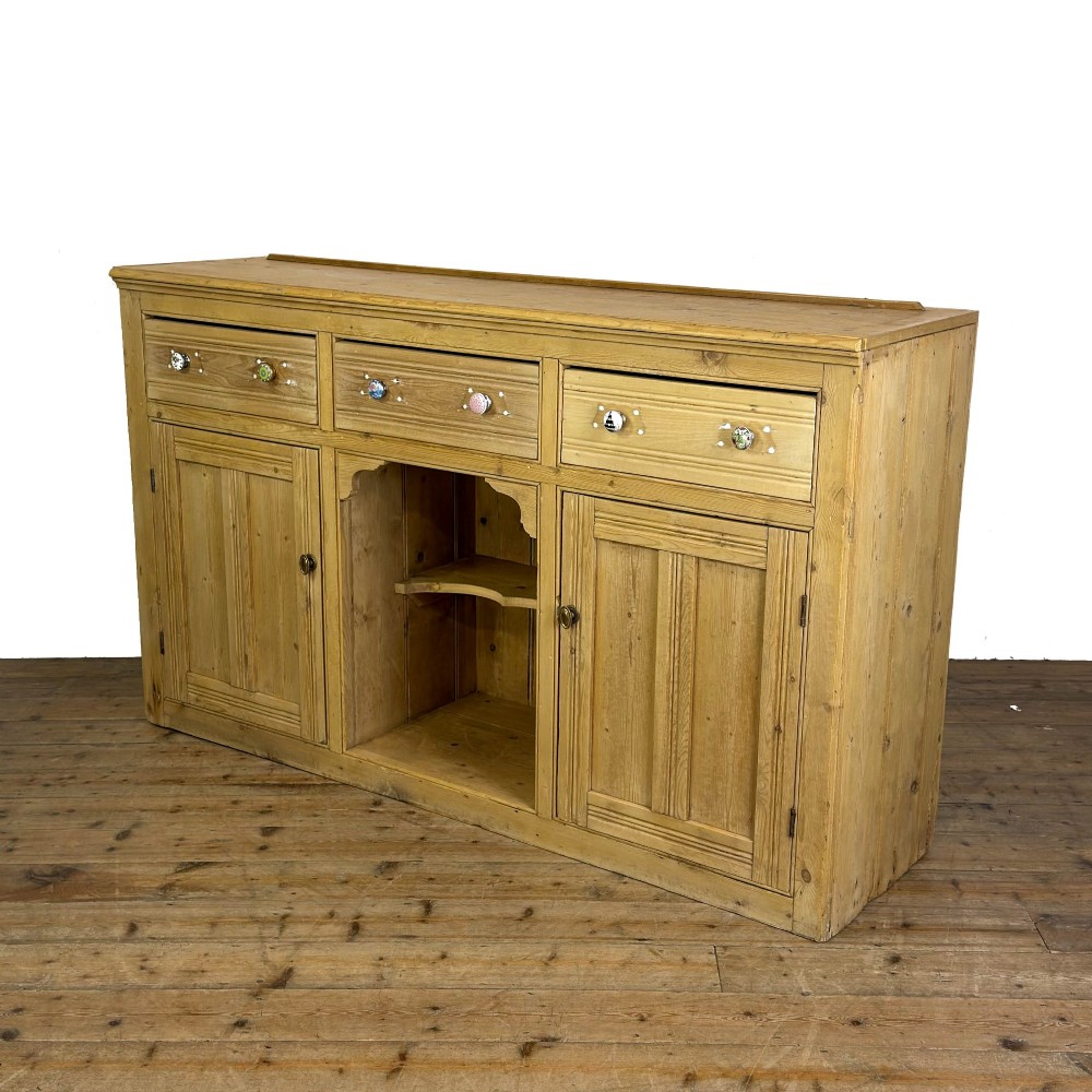 A Victorian pine dresser base with three frieze drawers, ceramic drawer knobs, two panelled cupboard doors, and a central open shelving recess. View II