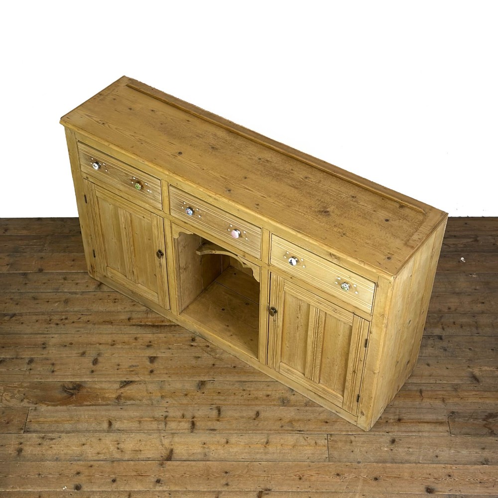 A Victorian pine dresser base with three frieze drawers, ceramic drawer knobs, two panelled cupboard doors, and a central open shelving recess. View III