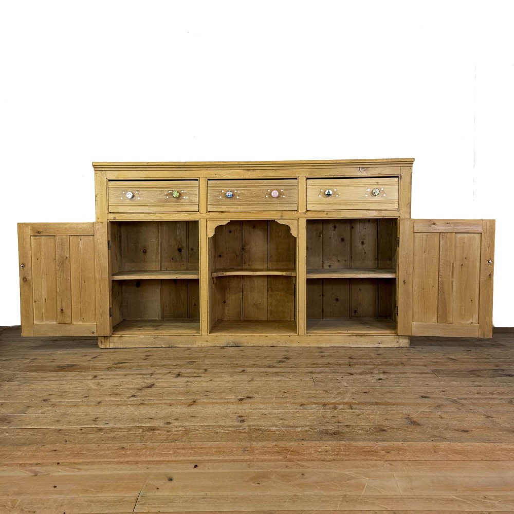 A Victorian pine dresser base with three frieze drawers, ceramic drawer knobs, two panelled cupboard doors, and a central open shelving recess. View IV