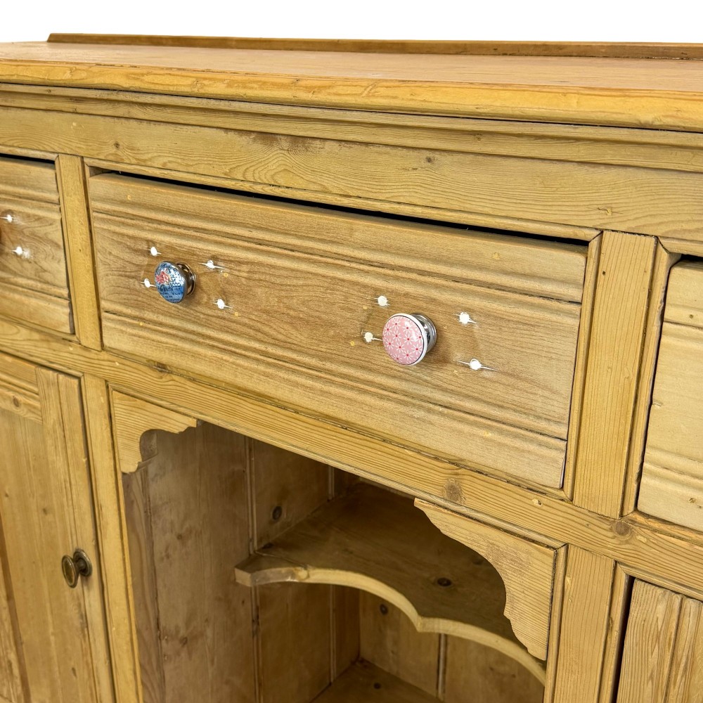 A Victorian pine dresser base with three frieze drawers, ceramic drawer knobs, two panelled cupboard doors, and a central open shelving recess. View V