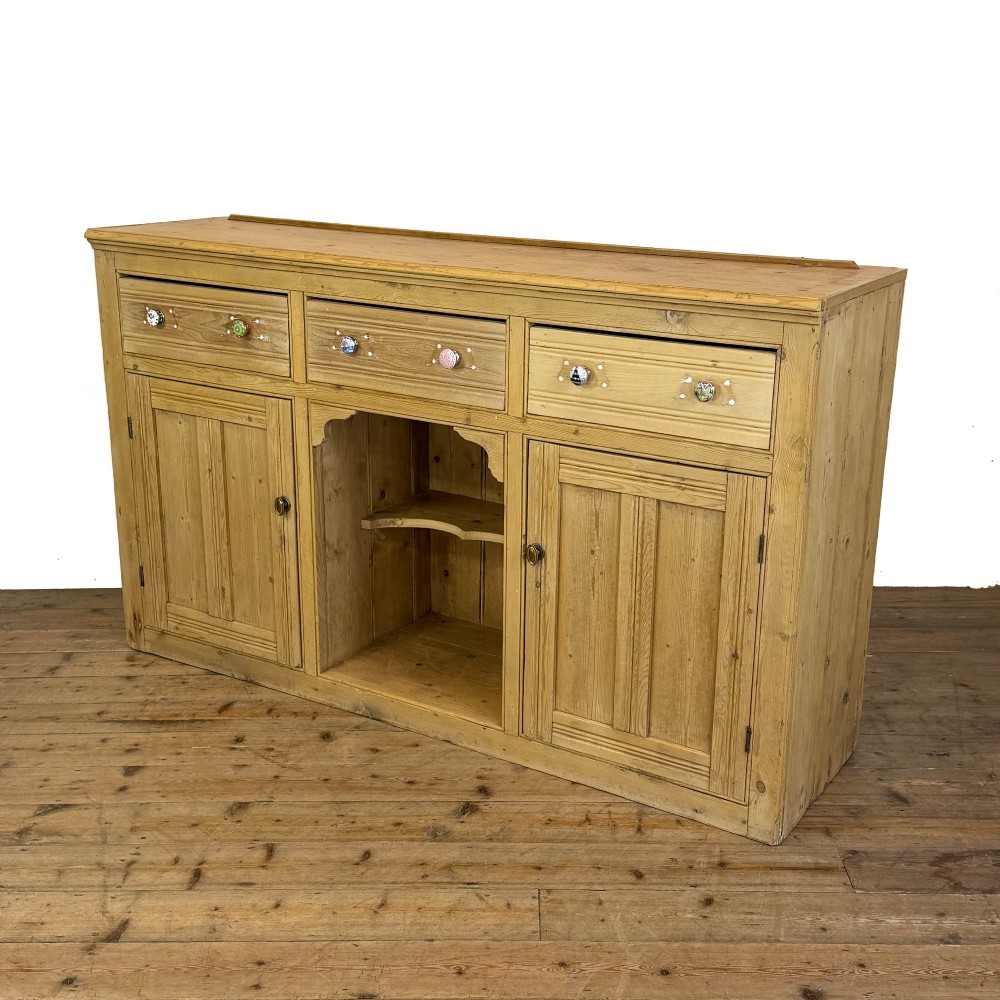 A Victorian pine dresser base with three frieze drawers, ceramic drawer knobs, two panelled cupboard doors, and a central open shelving recess. View XI