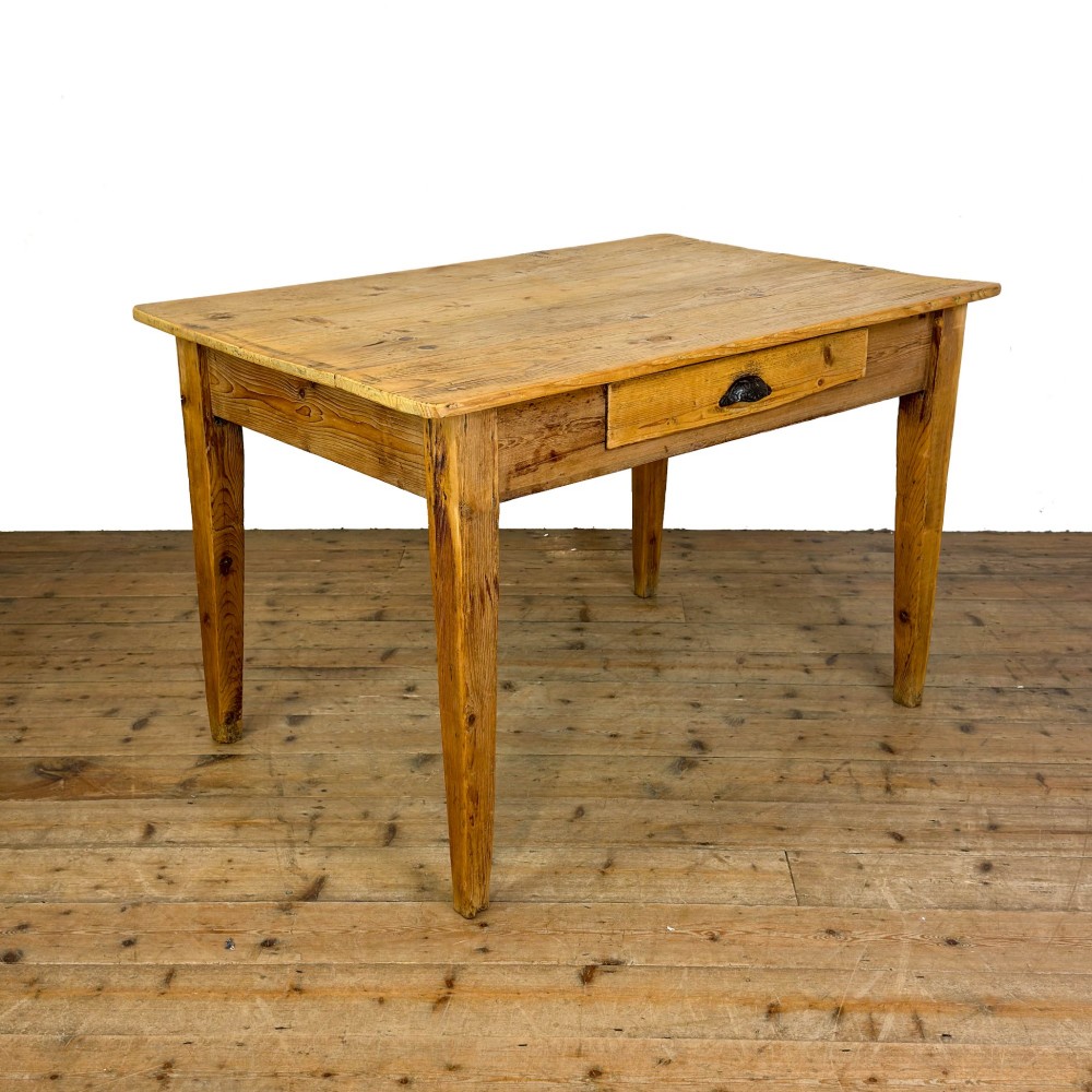 An antique pine kitchen table featuring a plank top, a single apron drawer with a cast iron cup handle, and four tapered square legs.