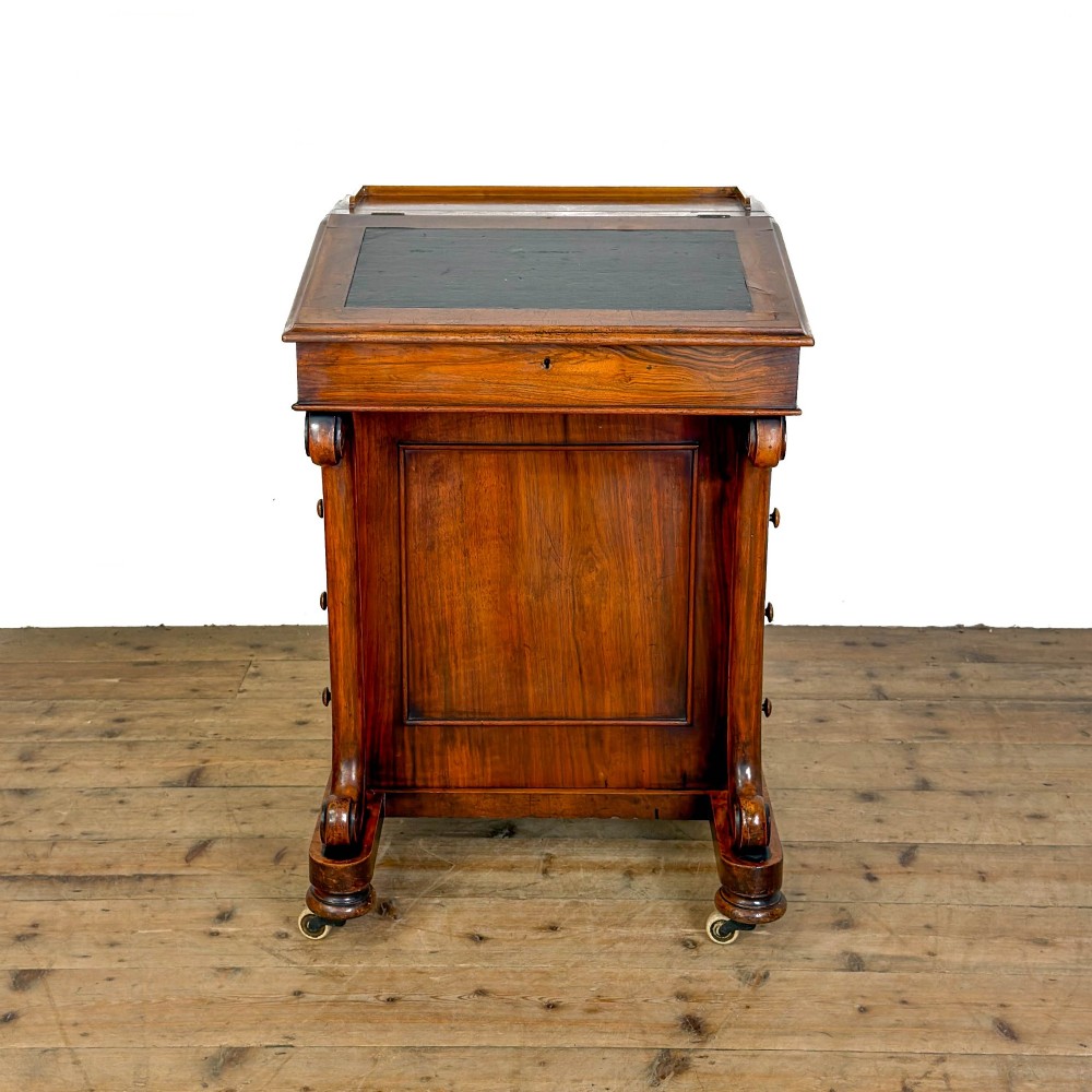 A Victorian walnut davenport desk featuring a slanted dark leather writing surface, carved front scroll supports, side drawers, and brass castors.