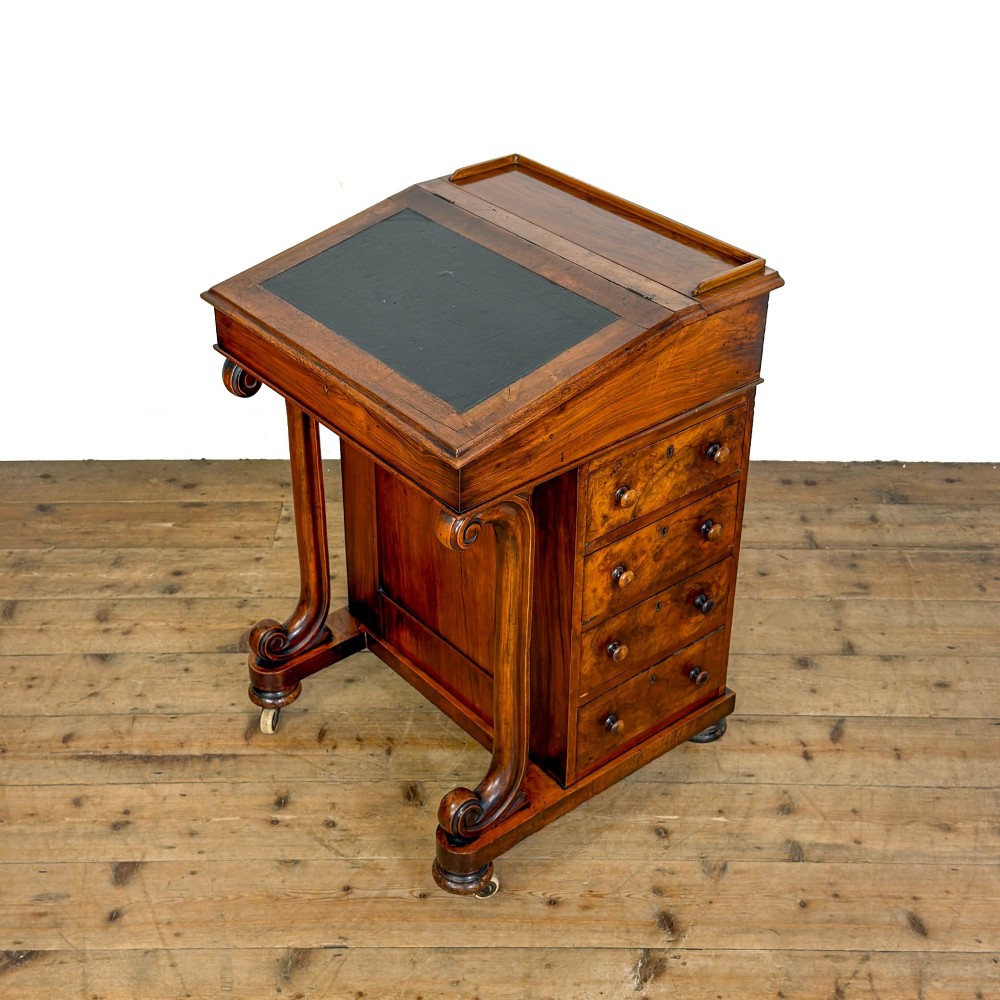 A Victorian walnut davenport desk featuring a slanted dark leather writing surface, carved front scroll supports, side drawers, and brass castors. View III