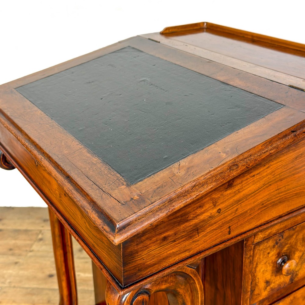A Victorian walnut davenport desk featuring a slanted dark leather writing surface, carved front scroll supports, side drawers, and brass castors. View V