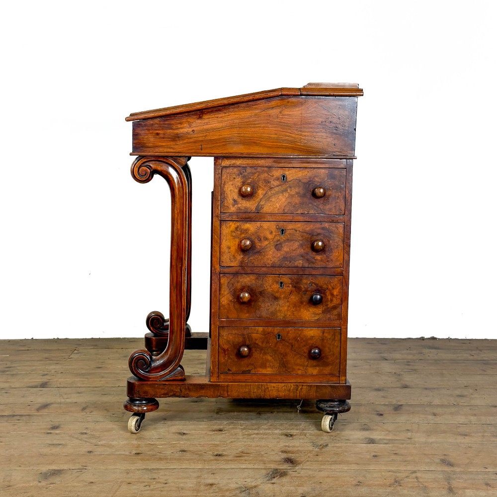A Victorian walnut davenport desk featuring a slanted dark leather writing surface, carved front scroll supports, side drawers, and brass castors. View VII