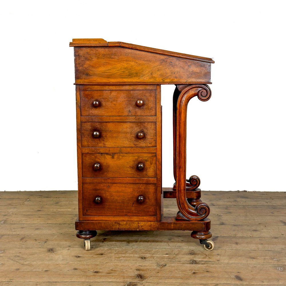 A Victorian walnut davenport desk featuring a slanted dark leather writing surface, carved front scroll supports, side drawers, and brass castors. View IX