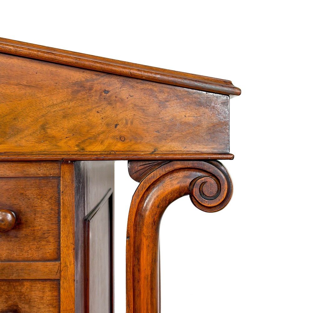 A Victorian walnut davenport desk featuring a slanted dark leather writing surface, carved front scroll supports, side drawers, and brass castors. View X