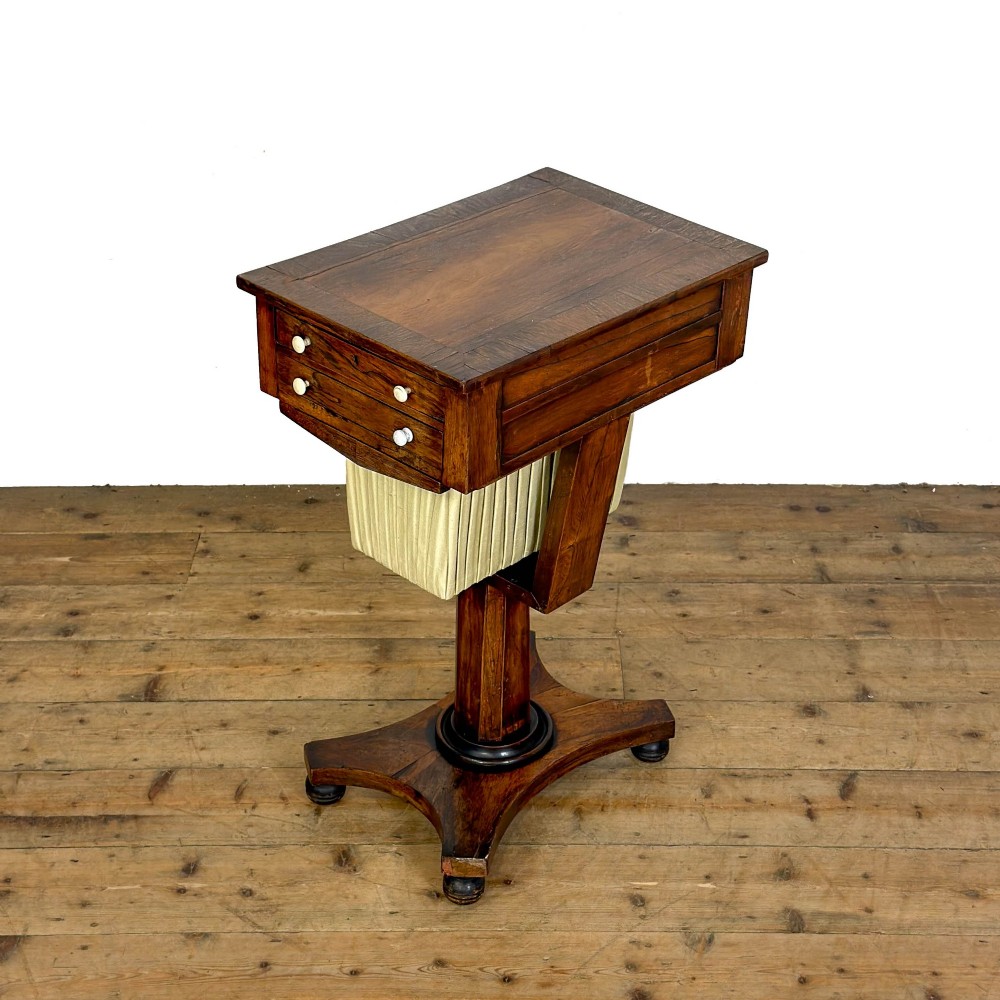 A 19th-century rosewood work table featuring small drawers, a pleated cream fabric basket underneath, a central pedestal support, and a quadruped base.