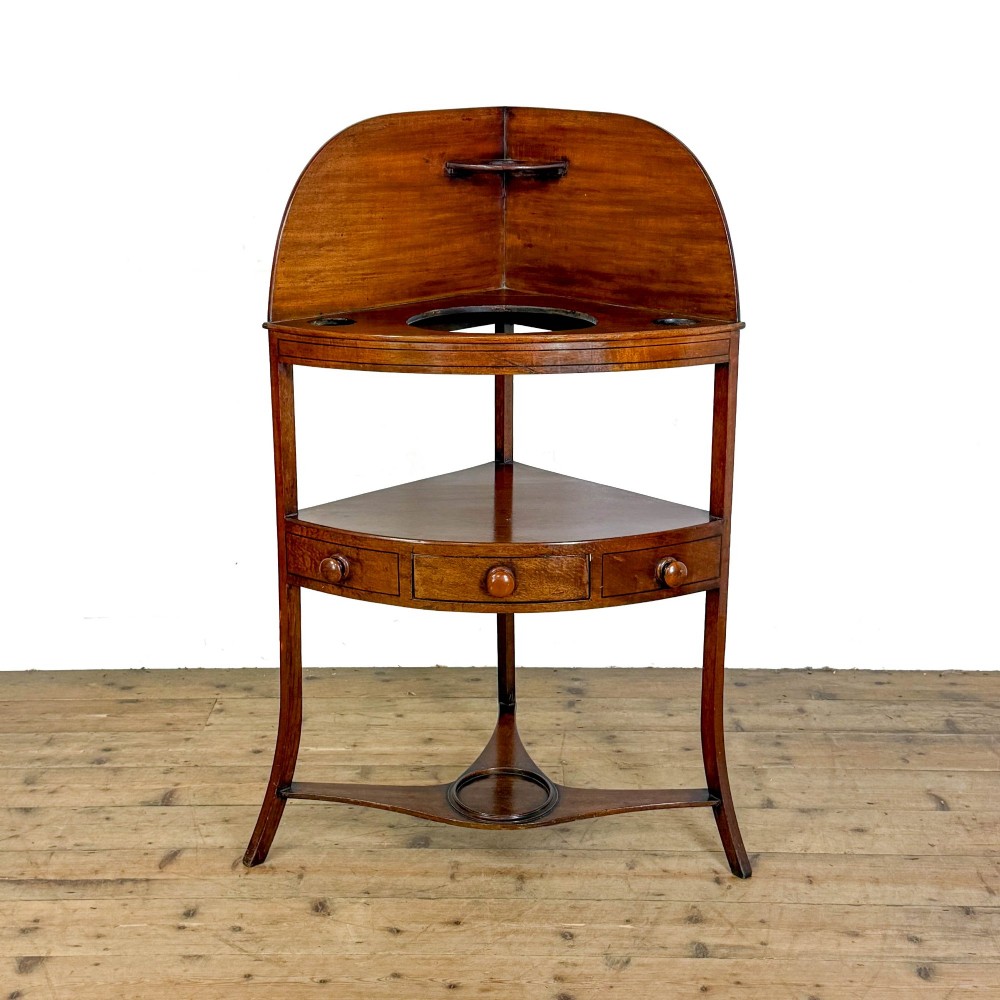 An antique mahogany corner washstand featuring a curved splashback, top circular basin cut-outs, a central drawer, and splayed legs connected by a lower stretcher shelf.