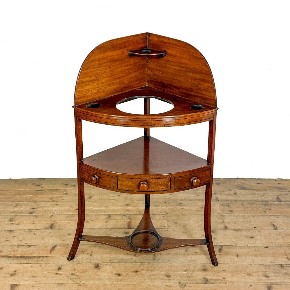 An antique mahogany corner washstand featuring a curved splashback, top circular basin cut-outs, a central drawer, and splayed legs connected by a lower stretcher shelf. View III