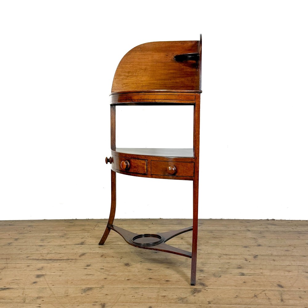 An antique mahogany corner washstand featuring a curved splashback, top circular basin cut-outs, a central drawer, and splayed legs connected by a lower stretcher shelf. View V