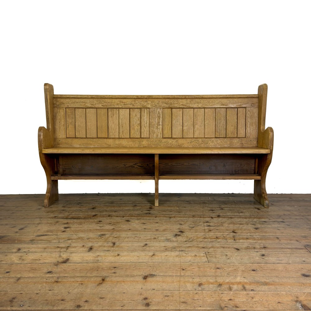 An antique pine chapel pew featuring a panelled backrest, integrated under-seat storage shelf, and sculpted bench ends.