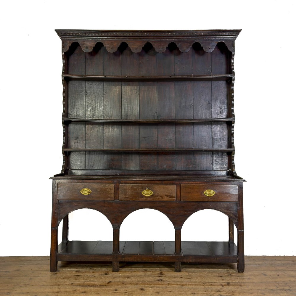 A dark oak 19th-century Welsh dresser featuring a three-tier open plate rack with a scalloped canopy above three drawers and an open pot board lower section.