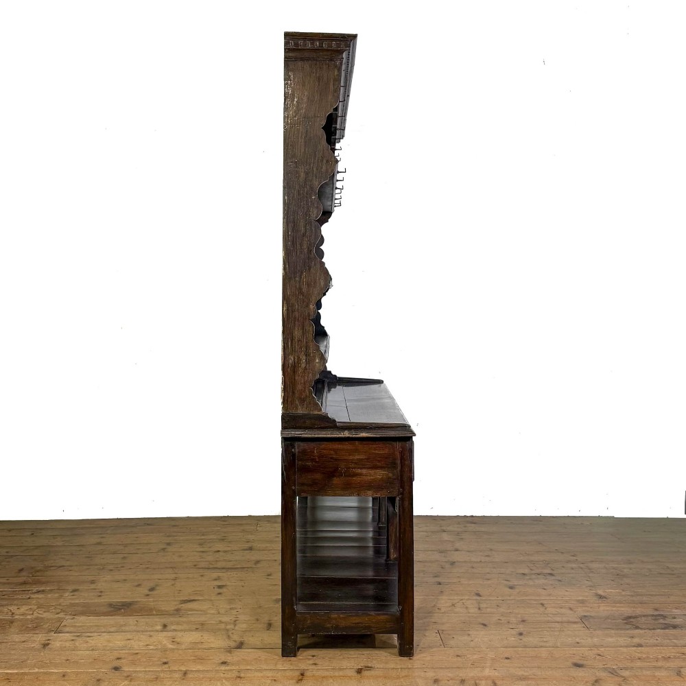 A dark oak 19th-century Welsh dresser featuring a three-tier open plate rack with a scalloped canopy above three drawers and an open pot board lower section. View VI